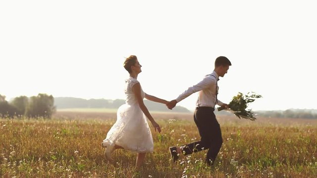 The newlyweds are running in the sunny field. The bride is throwing the black hat while the groom is holding the wedding bouquet.