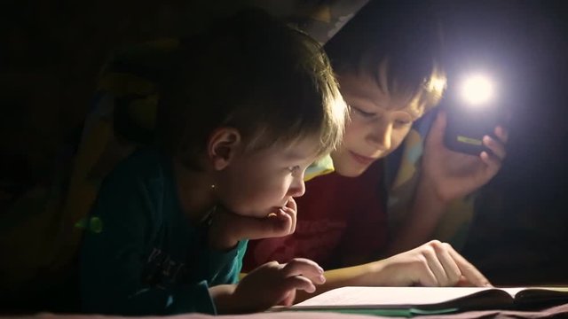 Children read a book under a blanket with a flashlight