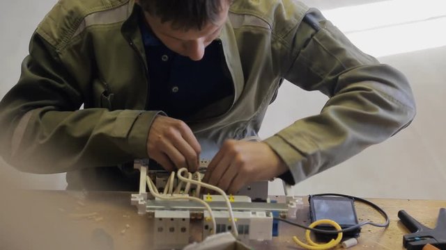 Professional electrician connects a safety switchgear using crimp. Man must finish connection of wires, then continue installation operation. Device case is located before him on wooden table beside