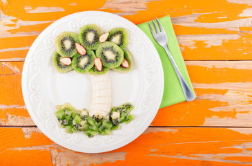 Fruit dessert for kids with kiwi, banana and strawberry on wooden background. Palm tree from fruit - original snack for party