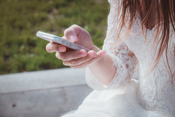 Tender bride in white dress with lace is holding phone in her hands. Beauty model girl in wedding day. Hands close-up in the park