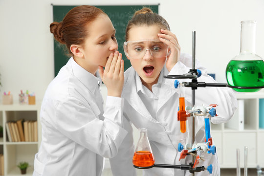Two school girls whispering and making experiment in chemistry class