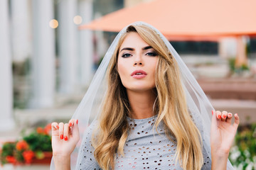 Portrait attractive girl with long blonde hair on street. She holds blue veil covering her head, looking to camera