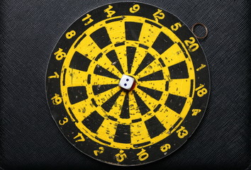 Used and vintage dartboard with arrow and dice in the scene appear a lot of damaged hole from arrow marked put on the black color leather surface as a background represent the entertainment game.