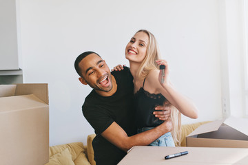 Couple exited about moving in new home, new life. Girl holding keys of new house,  boxes around, man and woman hugging, wearing black top and t-shirt Bright room with white wall on background.