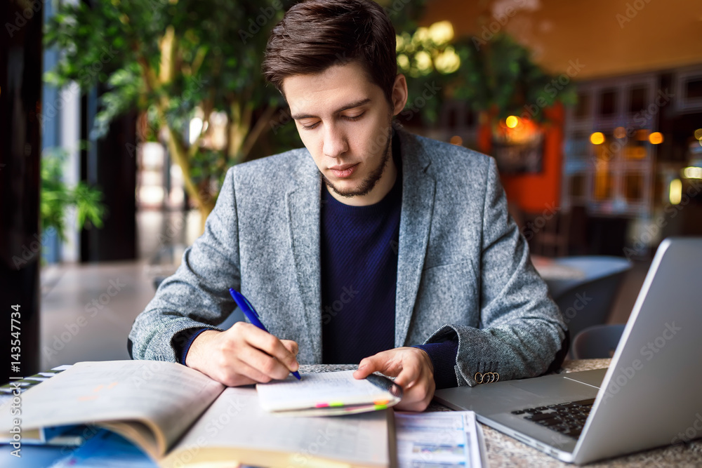 Wall mural Shot of young male  student sitting at table and writing on notebook. Young male student studying in cafe 
