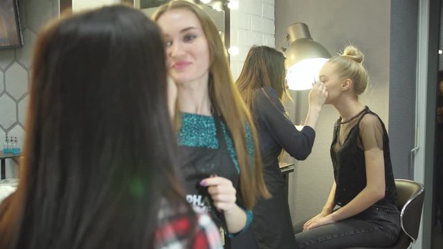 The work of the beauty salon in the evening, make-up artist adjusts eyebrows tweezers to beautiful brunette girl in checkered red shirt, next stylist putting makeup for pretty girls blonde.