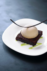 Plated dessert: dark chocolate brownie with praline mousse and pistachio cream, on white plate and dark background.