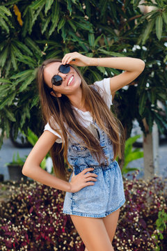 Cute slim tanned girl standing near tree. She wears white t-shirt, denim shorts and black sunglasses. She has long dark straight hair. She smiles and touches her sunglasses with her hand.