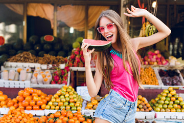 Brightful girl with long hair  on market with tropical fruits background. She is going to taste a slice of watermelon in hand.