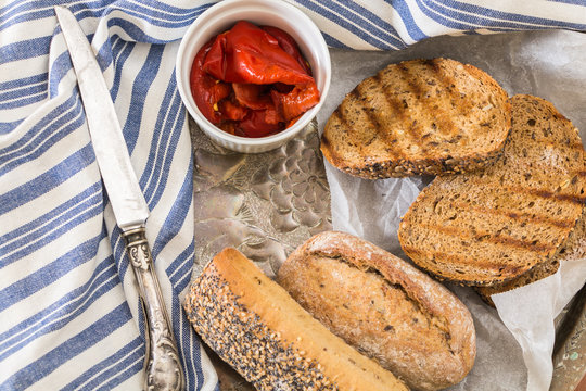 Whole wheat bread buns and grilled  bread slices, with roasted red peppers in bowl and knife.