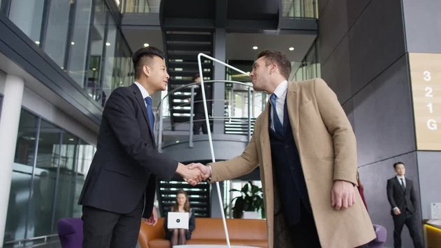  Businessmen meet & shake hands in lobby area of large modern office building