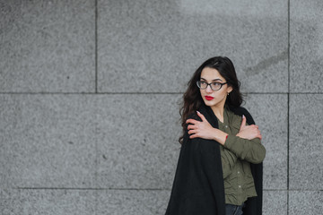 Fashion consept: beatiful young girl with long hair, glasses, red lips standing near modern wall wearing in green suit and grey jeans. She wanted talking on phone close eyes