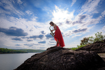 Beautiful woman wear red evening dress sound saxophone stand on the rocks over mountains and rivers...