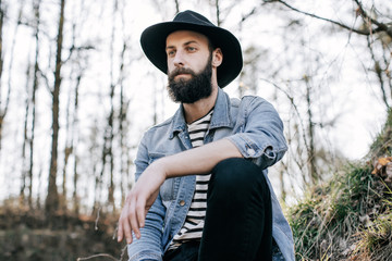 handsome bearded hipster man in a hat and denim jacket in the woods. Street style