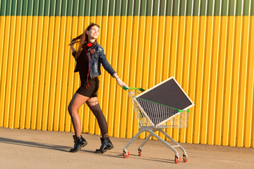 Cheerful stylish young girl in jeans jacket, shoes and skirt, listening to music on headphones with phone, on sunny day with shopping cart at supermarket, with chalk blackboard. space for text