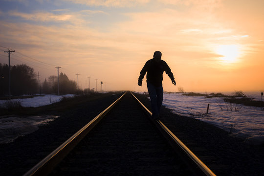 horizontal silhouette image of a man walking on the steel rail on the railroad track with a beautiful sunset in the background in early winter with room for text.
