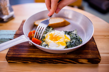Hand fork pricks tomato, breakfast consists of fried spinach, scrambled eggs, two slices of bread