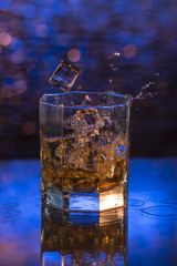 glass of whisky with dropped ice and splashes on a blue bokeh background