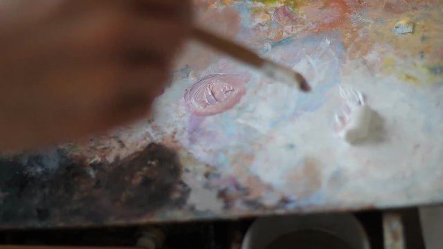 Mixing paint on palette, close-up. a painter with a long brush made of natural cloth blends paint white red and pink flowers. it turns a delicate peach shade for painting the skin.