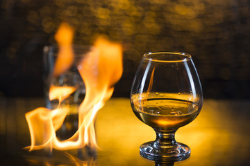 Glass o whisky with ice and wineglass of coghac and fire flames on yellow bokeh background