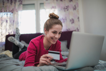 Young woman holding credit card and shopping online in bedroom alone