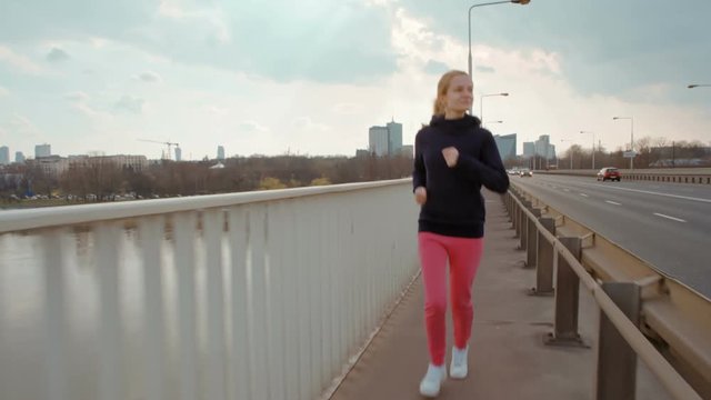 Attractive Young Woman in Sportswear is Running on a Bridge on the background of the Modern City