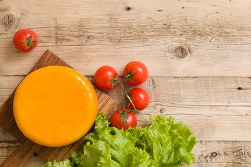 Top view on big cheese head in yellow vacuum package on wooden board served with tomatoes and fresh salad. Serving French homemade cheese. Food concept