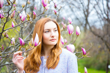 Beautiful red-haired young woman standing near blooming magnolia branches in spring garden