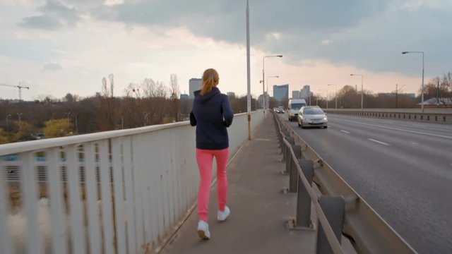 SlowMotion of Nice Young Woman in Sportswear Running on a Bridge in the City. Female jogging back view