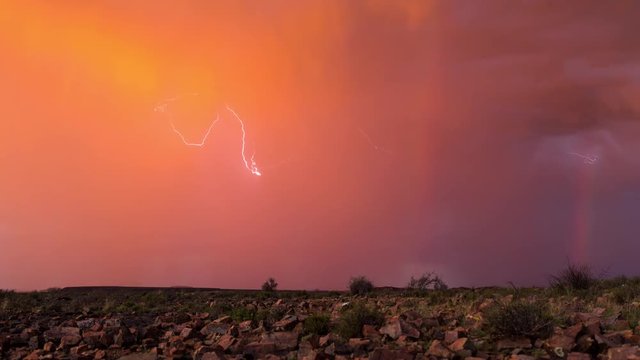 A static timelapse of a rocky landscape against a vivid orange and magenta stormy sky with lightning flashes and a rainbow while rain is pouring down