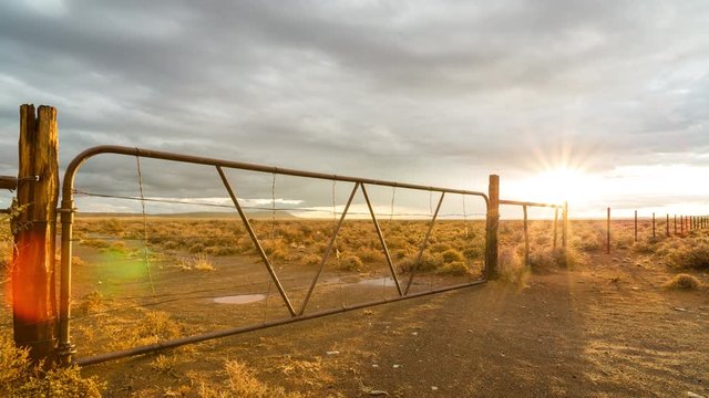 A beautiful static timelapse after a storm with a drenched Karoo landscape of an old rusted gate and fence with puddles of water while the sun dips through the clouds with an intense golden glow