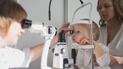 Doctor optometrist checks little girl's eyesight - mother and child in ophthalmologist room