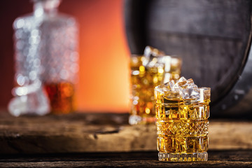 Whiskey. Two cups full of beverage whiskey brandy or cognac with ice cubes in retro style. Old oak barrel in the background.