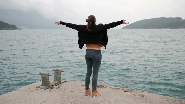 Woman stand on pier, raise her hand. View from back. Camera show slow motion of raising hands of brunette with long hair in black knitted sweater, grey slim jeans, orange boots and top under jacket
