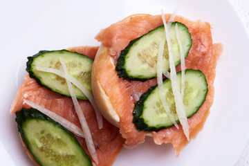 Bagel with salmon, cucumber, onion on white plate. Front view.