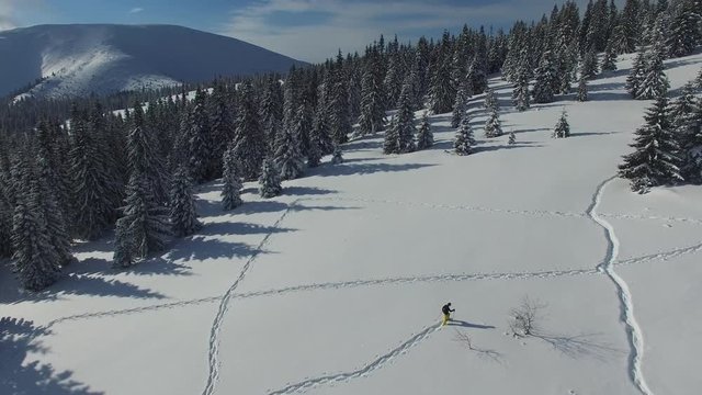 Aerial view of a traveler who is walking in snowshoes on a mountain slope, paving his way in deep snow, to his tent and reaching it happily waving his arms. Amazing  adventure in winter mountains.