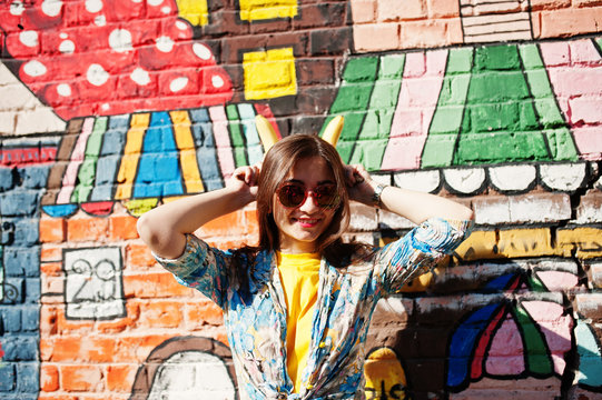 Beautiful fun teenage girl with bananas at hands, wear yellow t-shirt, jeans and sunglasses near graffiti wall.