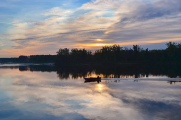 River at dawn and the silhouette of a fisherman in a boat in the distance. Reflection of clouds in water.