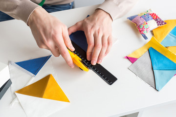 closeup on men hands of tailor, who is sitting behind a white desk and cut out colorful pieces of fabrics with rotary cutters for sewing patchwork items. Handwork and hobby concept.