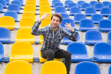 Attractive sporty young man model in blue shirt sitting on blue stadium seats after training staring at field