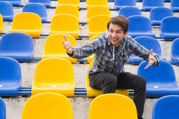 Attractive sporty young man model in blue shirt sitting on blue stadium seats after training staring at field