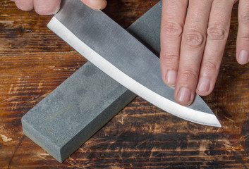 Knife sharpening. Hands holding knife and whetstone on the old wooden cutting board.