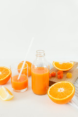 A bottle of orange fresh on a white table, next to a glass of fresh and oranges with lemon.