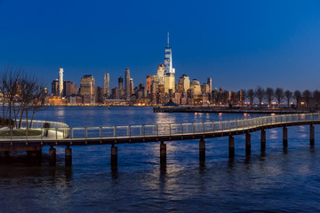 Fototapeta premium New York City Financial District skyscrapers at sunset and Hudson River from Hoboken promenade. Lower Manhattan skyline and pedestrian bridge from New Jersey