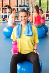 Beautiful young woman sitting on the blue pilates ball and smiling. Group of female athletes sitting behind.