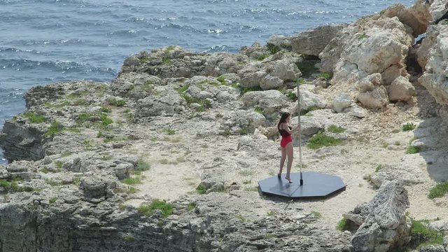 Pole dance fitness exercise on a rock at Cape Tarkhankut. Woman pole dancer in red during outdoor workout above the sea in summer. Long shot.