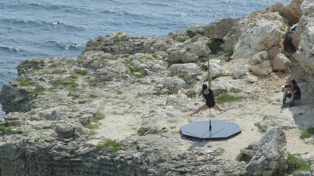 Photographer taking a photo of young woman pole dancer during pole dance fitness exercise on the edge of cliff