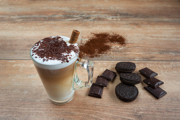 Cappuccino in glass cup with brown biscuits and pieces of chocolate on brown wooden board.