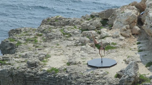 Outdoor pole dance fitness exercise above the sea. Fit girl pole dancer in bodysuit dancing on the edge of a cliff